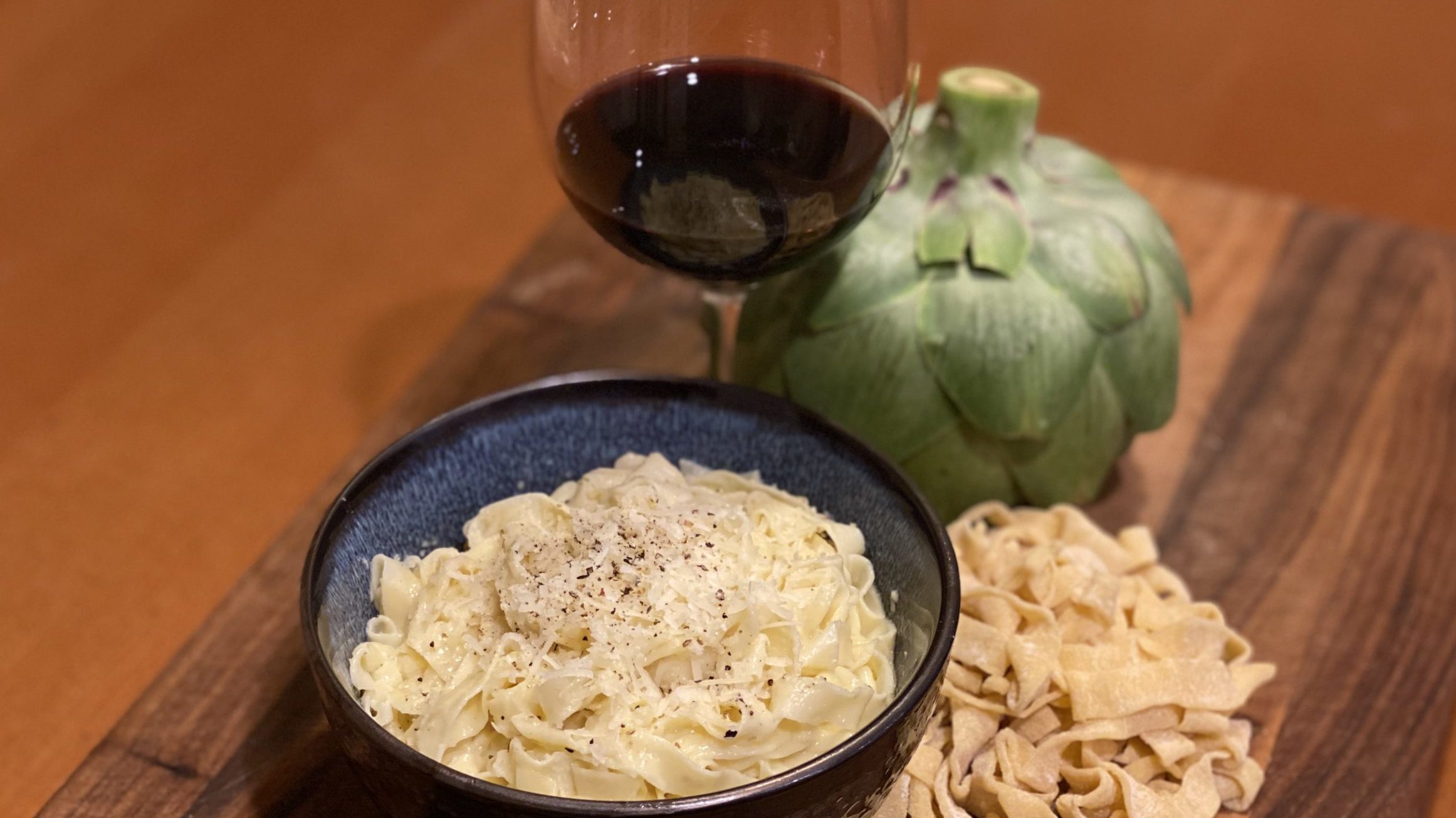 Bowl of fettuccini with alfredo sauce next to an artichoke and a glass of wine.