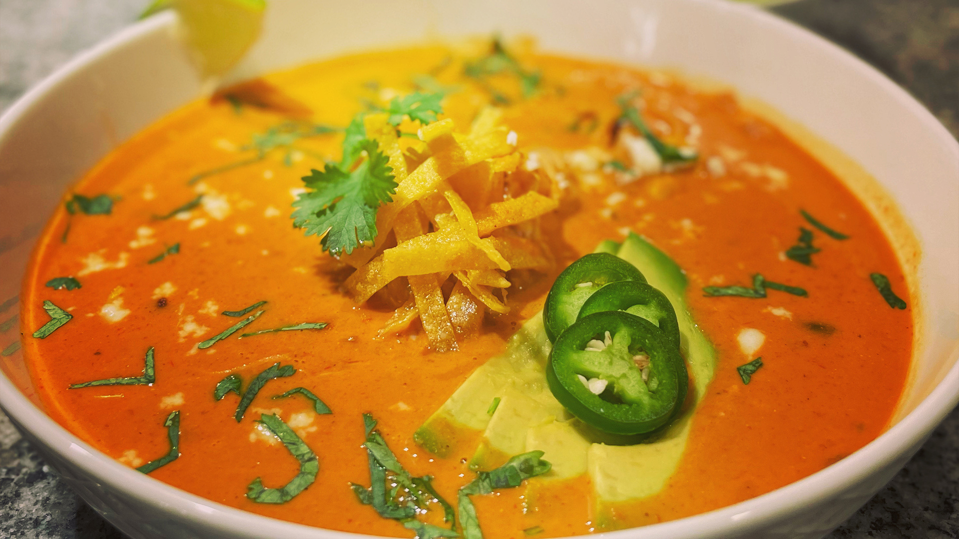 A bowl of chicken enchilada soup garnished with fried tortilla strips, avocado, and jalapeno peppers.